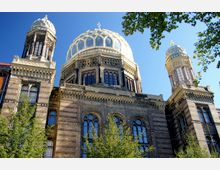 Backstein-Gebäude mit einer verzierten großen Kuppel und zwei kleineren Türmen, kombiniert mit Rundbogenfenstern und reichhaltigen architektonischen Details; im Vordergrund sind grüne Baumkronen sichtbar, der Himmel ist klar und blau.