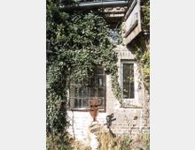 Eine alte Backsteinfassade, teils überwuchert mit Efeu, mit einem rechteckigen Metallfenster und einem kleineren Fenster mit verzierten Gittern. Darunter steht eine rostige metallische Skulptur, umgeben von etwas Gras und Wildpflanzen.