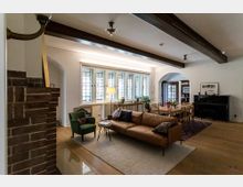 Wohnzimmer mit hellen Wänden und holzgetäfelter Decke, ausgestattet mit einem braunen Ledersofa, einem grünen Sessel und einem Klavier. Große Glasfenster mit Sprossen und dekorativer Bleiverglasung lassen Tageslicht herein, während ein Esstisch aus Holz mit sechs Stühlen sich neben einem großzügigen Teppich befindet. Im Vordergrund ist ein gemauerter Kamin sichtbar.