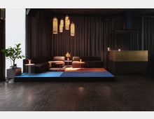 Gemütlicher Lounge-Bereich mit dunkelbraunen Sofas, kleinen runden Tischen auf farbigen Teppichen in Blau und Orange, weichen Vorhängen im Hintergrund sowie mehreren hängenden, warm beleuchteten Lampen. Rechts ist eine Theke mit goldener Verkleidung zu sehen, links steht eine Pflanze in einem mobilen Topf.