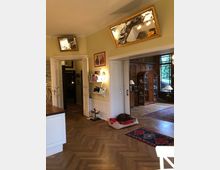 Wohnraum in einem Altbau mit gelb gestrichenen Wänden, Parkettboden und weiß lackierten Türen. Im Vordergrund ein Hundekorb auf dem Boden, darüber zwei schräg aufgehängte Spiegel, im Hintergrund ein Raum mit antikem Holzschrank und großen Fenstern, die auf einen grünen Außenbereich blicken.