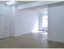 Ein leerer Raum mit weißen, strukturierten Wänden, einem Holzboden und einem großen Fenster, das natürliches Licht hereinscheint. Die Decke hat sichtbare Leuchtstofflampen, und links ist eine schlichte weiße Tür zu sehen.