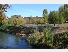 Industriegelände mit alten Backsteingebäuden im Hintergrund, umgeben von Bäumen und Grasflächen, im Vordergrund ein Fluss mit bewachsenen Ufern.
