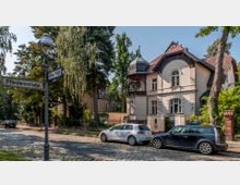 Eine historische Villa mit dekorativem Erker und einem kleinen Zwiebelturm in einem grünen, von Bäumen gesäumten Wohngebiet. Im Vordergrund sind zwei Straßenschilder (Pfleidererstraße und Geibelstraße) sowie zwei parkende Autos auf einer gepflasterten Straße zu sehen.