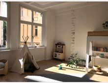 Helles Kinderzimmer mit großen Fenstern, durch die Tageslicht einfällt. Es sind ein Tipi-Zelt, ein Hochbett mit eingebautem Regal, eine Spielzeugküche, ein Bücherregal sowie Pflanzen und Spielsachen zu sehen.