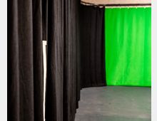 Ein schmaler Raum mit schwarzen Vorhängen an der linken Wand und einem leuchtend grünen Hintergrundvorhang auf der rechten Seite. Der Boden ist grau und die Decke hat eine schlichte, technische Ausstrahlung.