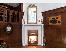 Gemütliches Wohnzimmer mit einem weißen Kamin in der Mitte, darauf ein großer goldener Spiegel und dekorative Elemente. Der Raum ist durch dunkle Holzvertäfelungen, eine antike Wanduhr, ein abstraktes Gemälde und ein Bücherregal geprägt.