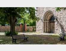 Historische Kirche mit romanischem Portal aus Stein, eingerahmt von verzierten Säulen und einem Holztor, umgeben von einer grünen Baumlandschaft und einer niedrigen Steinmauer; im Vordergrund stehen zwei Parkbänke.