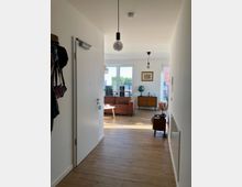 Flur in einer modernen Wohnung mit hellem Holzfußboden, einer offenen Tür, die zu einem Wohnbereich führt. Im Hintergrund sind ein braunes Sofa, ein runder Couchtisch, ein Globus und eine Kommode vor Fenstern mit Blick nach draußen sichtbar.