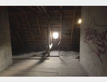 Dunkler Dachboden mit sichtbarer Holzbalkenkonstruktion und einem kleinen Fenster, das Licht hereinscheinen lässt. Rechts an der Wand ist ein großes, lilafarbenes Graffiti zu sehen.