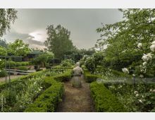 Ein symmetrischer Garten mit gepflegten Buchsbaumhecken, blühenden Rosensträuchern und einer steinernen Büste im Zentrum, umgeben von Bäumen und Laubpflanzen unter einem bewölkten Himmel. Im Hintergrund ist eine kleine, überdachte Gartenlaube erkennbar.