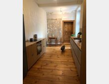 Schmale Küche mit Holzboden, links und rechts moderne Holzeinbauschränke mit Arbeitsflächen. Im Hintergrund ein runder Holztisch mit zwei Stühlen vor einer grob verputzten Wand und einer hölzernen Tür; eine Katze läuft über den Boden.