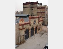 Backsteinbau mit historischer Fassade und halbkreisförmigen Fensterbögen aus Eisen sowie Zierelementen aus roten Ziegeln. Im Vordergrund ist ein gepflasterter Hof sichtbar, im Hintergrund ergänzen umliegende Gebäude die Szene.