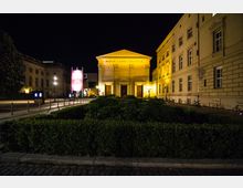 Nachtaufnahme des Maxim Gorki Theaters in Berlin, einem klassizistischen Gebäude mit Säulenfassade und Dreiecksgiebel. Im Vordergrund befindet sich eine gepflegte Hecke, und die Umgebung ist von beleuchteten Straßenlaternen und historischen Gebäuden eingerahmt.