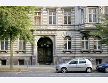 Fassade eines älteren, grauen Wohngebäudes mit verzierten Fenstern und einem großen Eingang mit Rundbogen. Vor dem Gebäude stehen zwei Bäume sowie geparkte Autos an der Straße; die untere Hauswand ist mit Graffiti bedeckt.