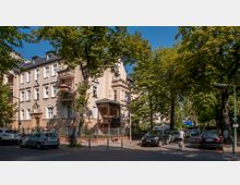 Straßenszene mit einem historischen Wohnhaus im Gründerzeitstil, umgeben von großen Bäumen. Parkende Autos säumen die Straße, und eine Person fährt mit einem Fahrrad auf der rechten Seite.