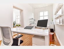 Helles, modernes Büro mit Holzboden, weißen Wänden und großen Fenstern. Im Vordergrund steht ein weißer Schreibtisch mit Computer und Dekoration, im Hintergrund sind Regale, ein weiterer Arbeitsplatz und Pflanzen zu sehen.