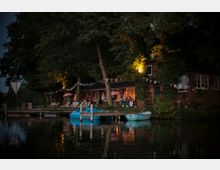 Ein kleiner Biergarten mit Lichterdekoration und bunten Wimpeln befindet sich unter Bäumen an einem ruhigen Ufer. Im Vordergrund liegen blaue Boote an einem Holzsteg, während Gäste an Tischen und Liegestühlen beim Gebäude sitzen.