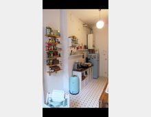 Kleine Küche mit hellen Wänden und gemustertem Boden. An der linken Wand befinden sich offene Regale mit Lebensmitteln, rechts ist ein Küchenschrank mit Geschirr, Kaffeemaschine und Mini-Backofen zu sehen. Ein Kühlschrank mit aufgesetztem Boiler steht in der Ecke, beleuchtet von einer Hängelampe.