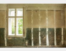 Innenraum eines renovierungsbedürftigen Zimmers mit weiß gerahmtem Doppelfenster, durch das Grün von Bäumen zu sehen ist. Die Wand zeigt abgeblätterten Putz und freiliegende, ungleichmäßige Farbschichten mit vertikalen Streifen und einem sichtbaren Steckdosenanschluss nahe dem Boden.