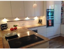 Moderne Küche mit glänzenden weißen Einbauschränken, einer Arbeitsplatte aus beigem Stein und Holzfußboden. Sichtbar sind ein Kochfeld, ein Doppelfenster-Backofen, eingelassene Deckenbeleuchtung sowie Küchendekoration wie Kürbisse und Kräuter.