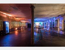 Weitläufiger Veranstaltungsraum mit hohen Decken, freigelegten Metallstützpfeilern und einer Mischung aus farbiger Beleuchtung in Blau- und Goldtönen. Der Raum verfügt über Stehtische mit Metallstühlen, eine Wand mit goldener Dekoration und einen Kühlschrank mit Getränken auf der linken Seite.