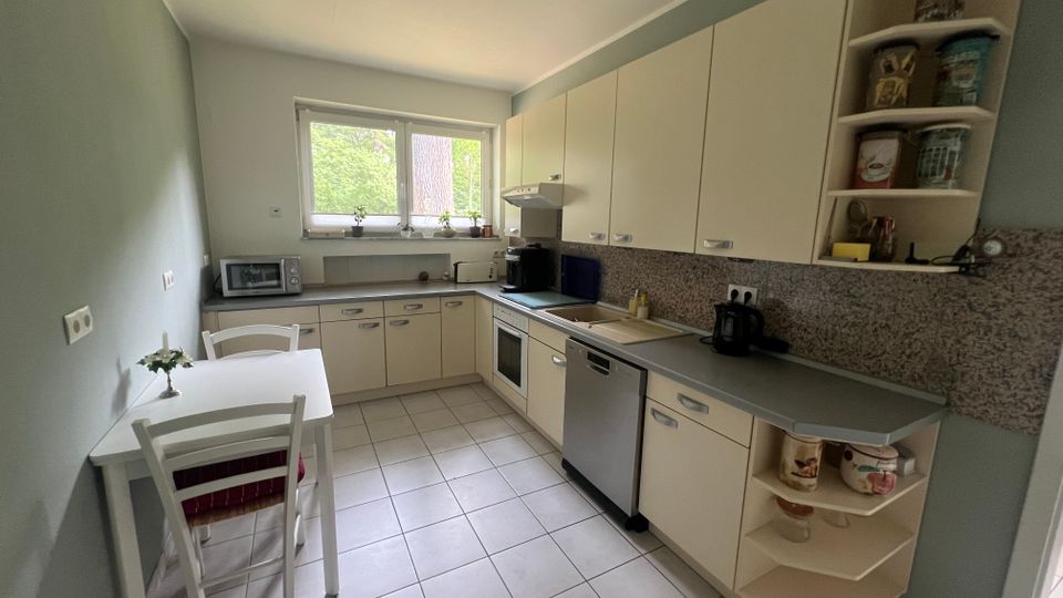 Helle Wohnküche mit L-förmiger Einbauküche in cremefarbenen Schränken, grauer Arbeitsplatte und Fliesenboden, mit Herd, Spüle und Geschirrspüler. Ein großes Fenster zur grünen Außenansicht sowie ein kleiner Esstisch mit zwei Stühlen stehen an der linken Wand.