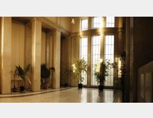 Heller Innenraum mit hohen, bodentiefen Fenstern, durch die weiches Licht einfällt. Der Raum ist mit cremefarbenem Marmor, Säulen und mehreren Topfpflanzen gestaltet.