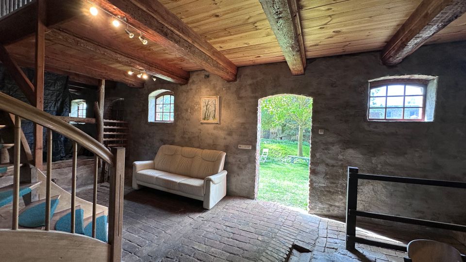 Rustikaler Innenraum eines umgebauten Scheunen- oder Stallgebäudes mit sichtbaren Holzbalken, Ziegelboden und verputzten Steinwänden. Eine offene Tür führt in einen grünen Garten; kleine Sprossenfenster und eine Holztreppe prägen den Raum.