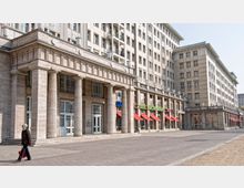 Straßenszene mit einem monumentalen Gebäude im neoklassizistischen Stil, geprägt von großen Säulen, hellen Steinfassaden und hohen Fenstern. Im Erdgeschoss befinden sich Geschäfte mit roten Markisen und bunten Schildern, während ein breiter Platz mit Pflastersteinen den Vordergrund einnimmt.