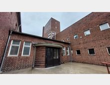 Backsteingebäude eines Stadtbades mit einem Eingang in der Mitte, gekennzeichnet durch eine schwarze Tür und ein rundes Vordach. Umgeben von schlichten Fenstern und hohen, roten Backsteinwänden.