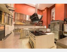 Gewerbliche Küche mit roten Wänden, weiß gefliester Rückwand und Edelstahlmöbeln. Sichtbar sind ein Herd mit Dunstabzug, Spülbecken, Hängeschränke, Regalflächen sowie ein großer Kühlschrank.