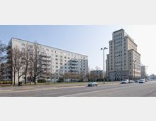 Zwei mehrstöckige Gebäude an einer breiten Straße, links ein länglicher Wohnblock mit Balkonen und rechts ein höheres Gebäude mit rechteckigem Grundriss und großen Fenstern. Im Vordergrund verläuft eine vierspurige Straße mit Autos, dahinter sind Gehwege, Rasenflächen und einzelne Bäume.