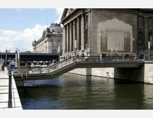 Historisches Gebäude mit Säulenfassade am Ufer eines Flusses, über dem eine Fußgängerbrücke mit Geländern verläuft. Im Hintergrund ist eine Kuppel und eine weitere Brücke sichtbar, während einige Personen die Brücke und das Uferbereich nutzen.