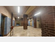 Ein Flur in einem Gebäude mit Backsteinpfeilern, hellem Fliesenboden und Wänden, die teils mit blauen Fliesen verkleidet sind. Im Hintergrund ein Durchgang mit einem Schild, auf dem "Alte Schwimmhalle" steht.