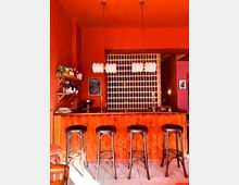 Interior of a small bar with vivid orange-red walls, a curved wooden counter, and four tall black bar stools on a tiled floor. Two pendant lights hang above the bar, with wall shelves of bottles and a chalkboard menu beside a white perforated partition behind the counter.