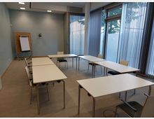 Kleiner Konferenzraum mit mehreren weißen Tischen und Stühlen, angeordnet in einer offenen U-Form. Im Hintergrund befindet sich ein Flipchart vor einer hellblauen Wand, während große Fenster mit vertikalen Jalousien den Raum erhellen.