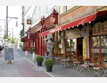 Straßenszene mit einem Restaurant mit arabischem Stil namens "Marrakesch" und rotem Vordach, dekoriert mit Laternen und Sitzplätzen im Freien mit Holztischen und Stühlen. Im Vordergrund stehen Topfpflanzen, und ein Holzschild wirbt für Cocktails; daneben verläuft ein Gehweg mit Bäumen und Straßenlaternen.