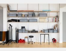 Offener Arbeitsbereich mit einem hohen Regal an der Wand, gefüllt mit grauen Kisten, Büromaterial, Büchern und Kunstutensilien. Unten befinden sich Hocker, Tische, Stative und rote Sandsäcke, auf einem hellen Holzboden.