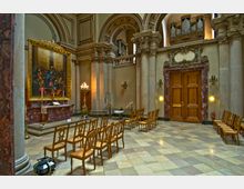 Innenraum einer historischen Kirche mit Marmorsäulen, verzierten Wänden und einer Orgel auf einer oberen Empore. Im Vordergrund stehen Holzstühle vor einem Altar mit einem großen Gemälde in einem goldenen Rahmen; daneben brennen Kerzen auf einem Kerzentisch.