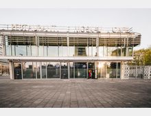 Modernes Gebäude mit großer Glasfassade und einem breiten Schriftzug „Restaurant Moskau Café“ auf dem Dach. Vor dem Eingang befinden sich eine gepflasterte Fläche und dekorative Zäune.