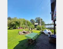 Gartenbereich am Seeufer mit Rasenfläche, Feuerstelle aus Steinen und Holzstangen, Liegestühlen und einem überdachten Sitzplatz mit Holzboden. Im Hintergrund Schilf, ein Bootshaus und ein See, umgeben von Bäumen und Sträuchern.