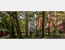 Backstein-Kirchengebäude mit neugotischen Elementen, umgeben von hohen Bäumen in einem parkartigen Garten. Ein moderner gläserner Aufzugsturm steht an der Seite des Gebäudes und fügt sich in die Umgebung ein.