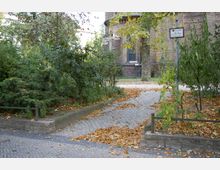 Gepflasterter Weg mit Herbstlaub, umgeben von Bäumen und Sträuchern, führt zu einem Gebäude aus rotem Backstein mit großen Bogenfenstern in einem parkähnlichen Bereich. Ein Schild steht am rechten Rand des Bildes.