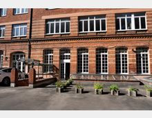 Innenhof vor einem mehrstöckigen Backsteingebäude mit großen, weiß gerahmten Fenstern und Rundbögen im Erdgeschoss. Im Vordergrund stehen Betonpflanzkübel, daneben befindet sich ein gläsernes Vordach und ein langes Glasdach über einem Lichtschacht.