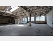 Großer, leerer Industriebereich mit hohen Decken, mehreren Fensterreihen aus Glasbausteinen und sichtbaren Rohrleitungen sowie Schienensystemen an der Decke. Der Boden ist aus Beton, und Licht fällt durch Oberlichter und Fenster.