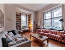 Wohnzimmer mit Holzboden und großen Fenstern, die viel Tageslicht hereinlassen. Der Raum ist mit einem grauen Sofa, einem gestreiften roten Sofa, einem Holztisch, Bücherregalen und einem Fernseher ausgestattet; Pflanzen und Dekorationselemente befinden sich auf der Fensterbank.