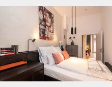 Modernes Schlafzimmer mit Doppelbett, weißen Wänden und dunklen Holzschränken; über dem Bett hängt ein großes Wandbild, daneben stehen Nachttischlampen. Zwei schwarze Pendelleuchten hängen von der Decke, und eine offene Tür führt in einen angrenzenden, hellen Raum mit Holzboden.