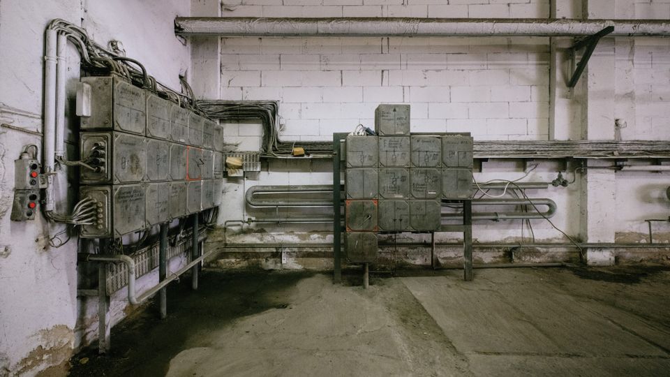 Innenansicht eines technischen Raums mit grauen Verteilerkästen und Elektronikinstallationen an einer weißen Ziegelwand. Der Boden ist aus Beton, und Kabel verlaufen entlang der Wand und zu den Kästen.