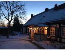 Fachwerkhaus in winterlicher Umgebung mit schneebedecktem Dach und beleuchteter Veranda, umgeben von einem schneebedeckten Garten und kahlen Bäumen bei Dämmerung.